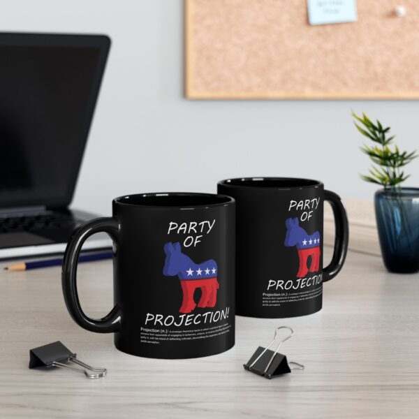 Two Party Of Projection black ceramic mugs with white text on table next to a laptop